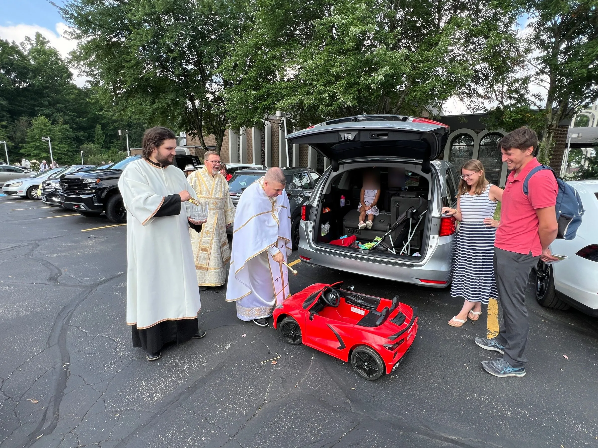 Clergy in white vestments blessing a family's minivan in the parking lot