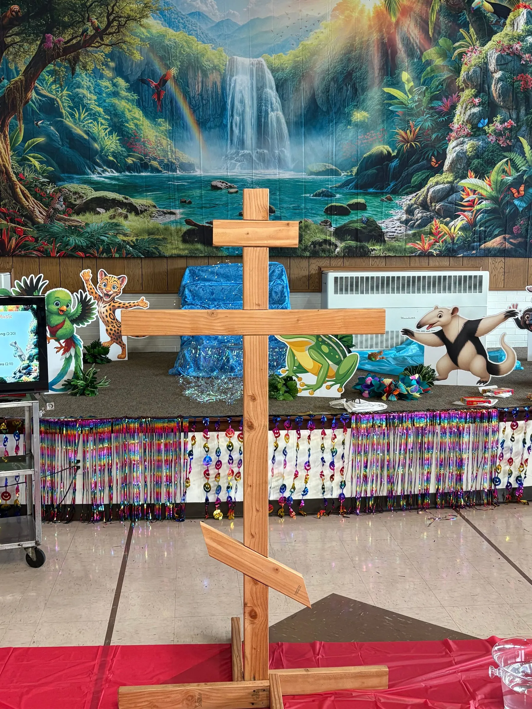 A wooden three-bar Byzantine cross standing before a painted rainforest waterfall