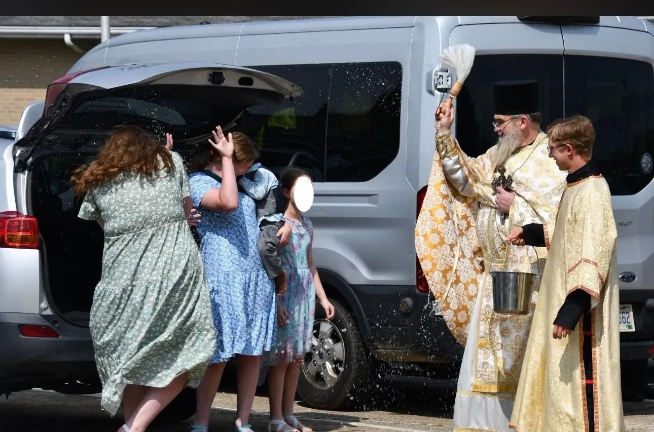 A priest sprinkling holy water over a laughing family beside their van