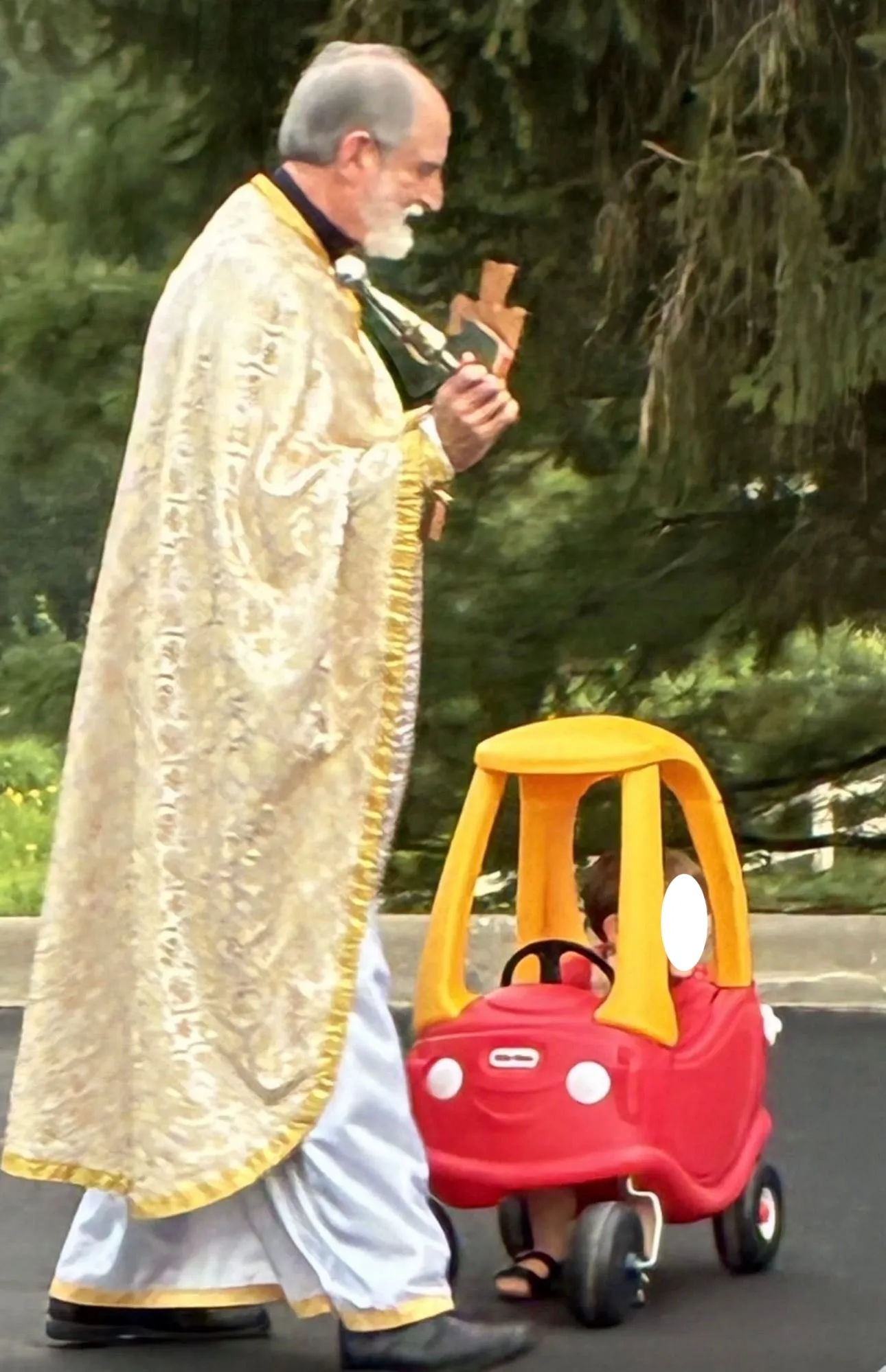 A priest in gold vestments walking past a child riding a toy Cozy Coupe car